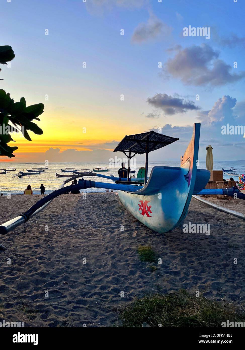 Tradizionale barca da pesca balinese con fuori di bordo che riposa sulla riva sabbiosa al tramonto a Jerman Beach, Kuta, Bali, Indonesia, il 12 aprile 2026. Foto Stock