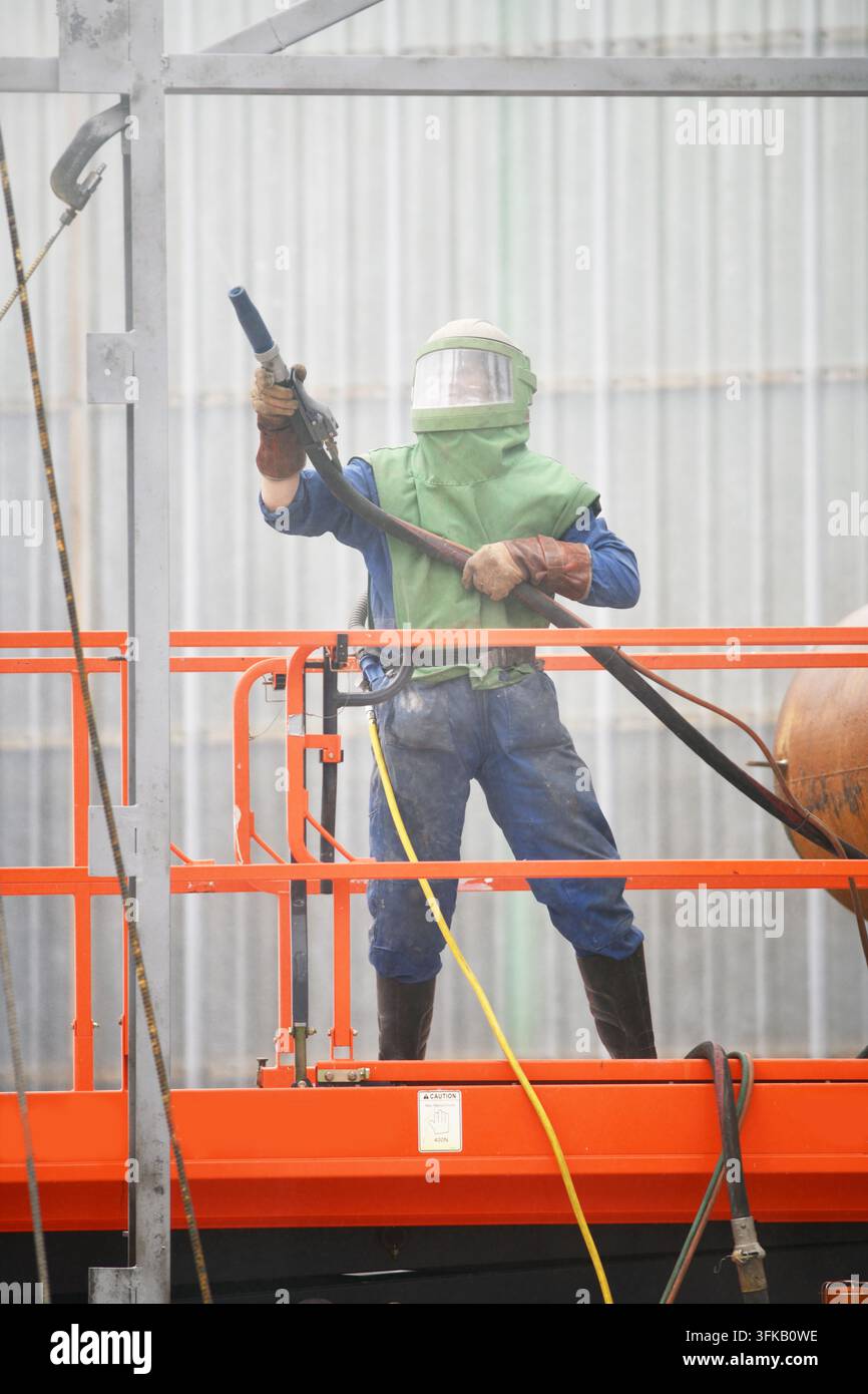 Un uomo in abiti protettivi aziona una sabbiatrice da una piattaforma a forbice per pulire le strutture in acciaio di un edificio danneggiato da un incendio, pronto per la ridipintura. Foto Stock