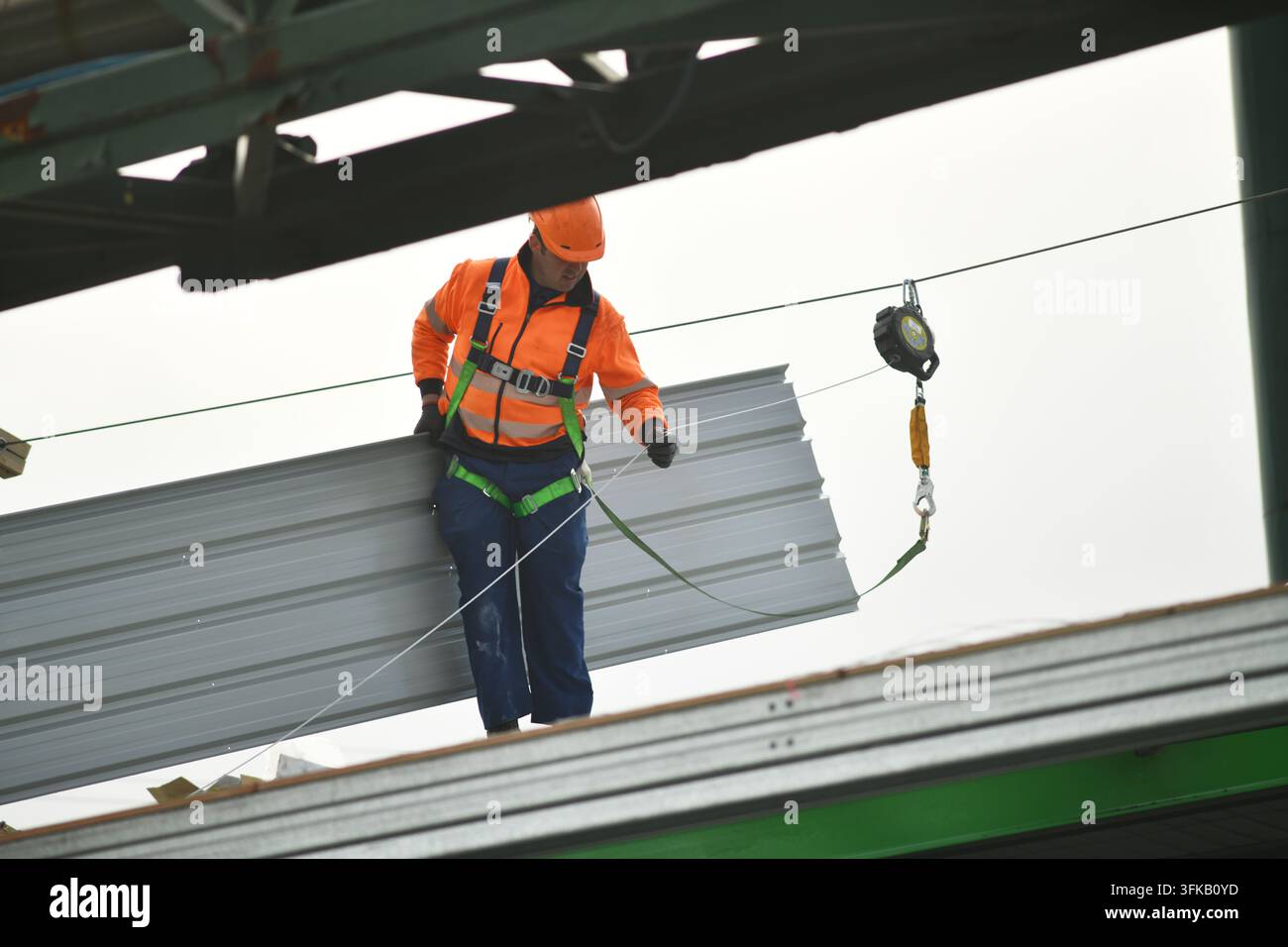 Un muratore che indossa un'imbracatura di sicurezza trasporta una lastra di ferro nuovo per il tetto al suo posto su un edificio commerciale. Foto Stock
