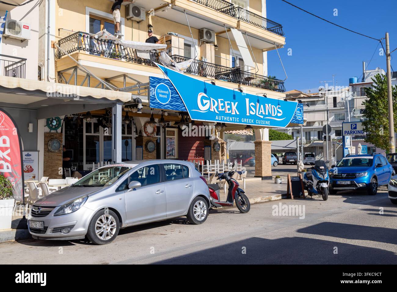Zante, Grecia, 1 settembre 2026: mostra un ristorante delle isole greche con posti a sedere all'aperto, auto parcheggiate, scooter e lavoratori su un balcone sopra il locale Foto Stock