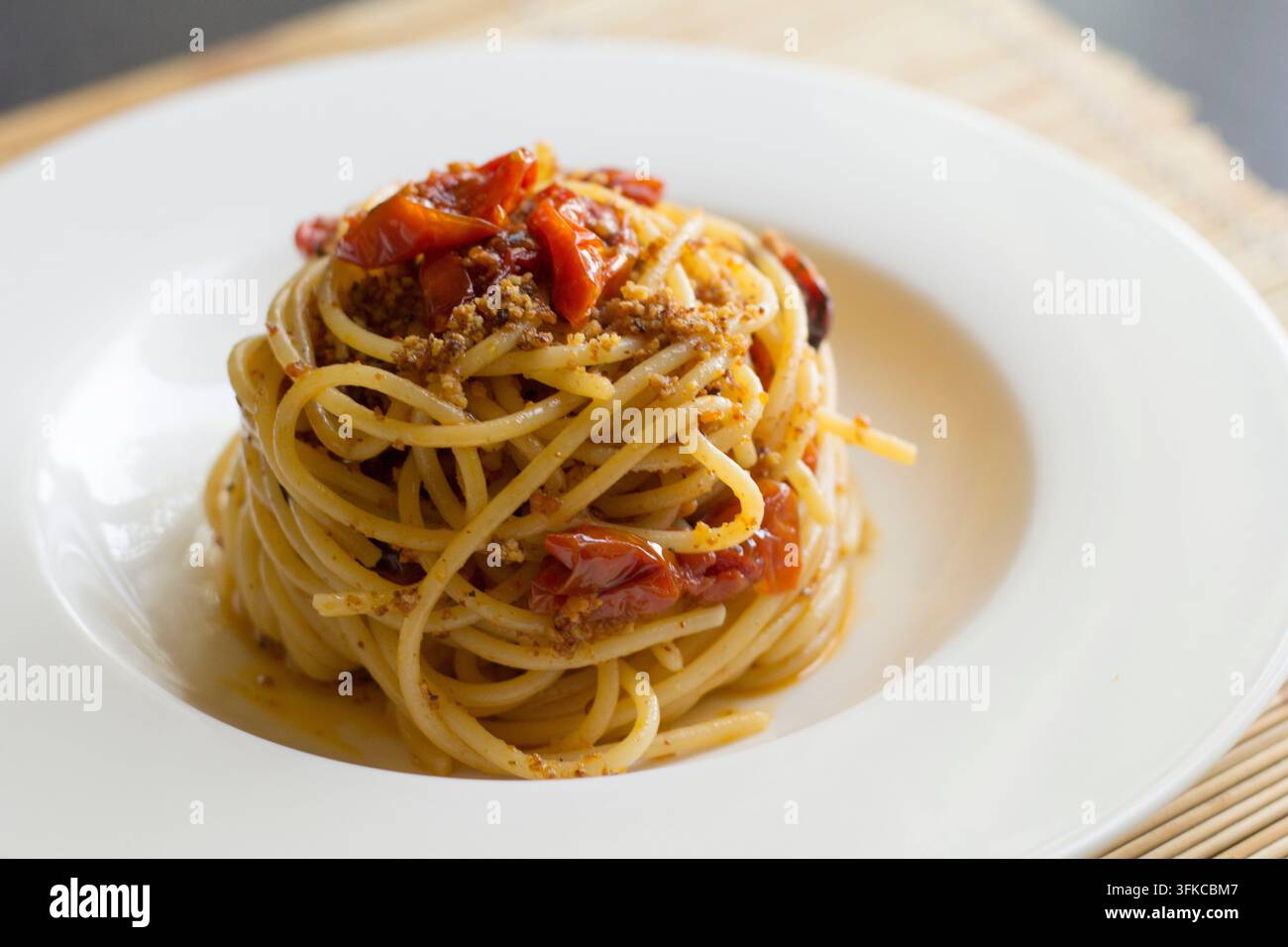 Spaghetti alti e attorcigliati con pomodorini confit e briciole di pane tostate su un piatto bianco, vista a 45 gradi, sfondo in stuoia di bambù Foto Stock