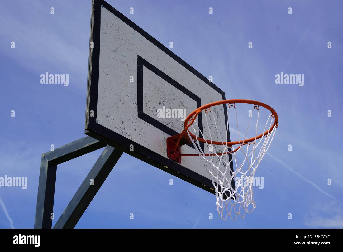 Un piano ad angolo basso che guarda verso un tabellone da basket all'aperto, il cerchio e la rete bianca montati su un palo di metallo contro un cielo azzurro con sottili strisce Foto Stock
