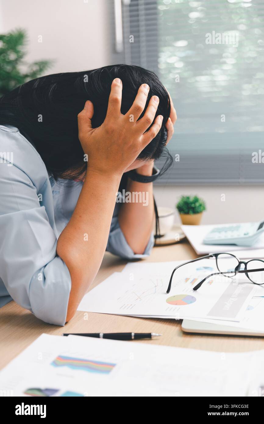 Donna d'affari sopraffatta che tiene la testa a una scrivania con laptop, calcolatrice e rapporti, mostrando burnout, stress lavorativo, pressione delle scadenze, w finanziaria Foto Stock