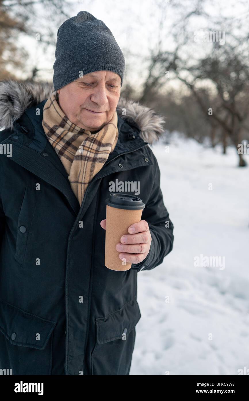 Uomo maturo che tiene una tazza di caffè da asporto in un parco innevato Foto Stock