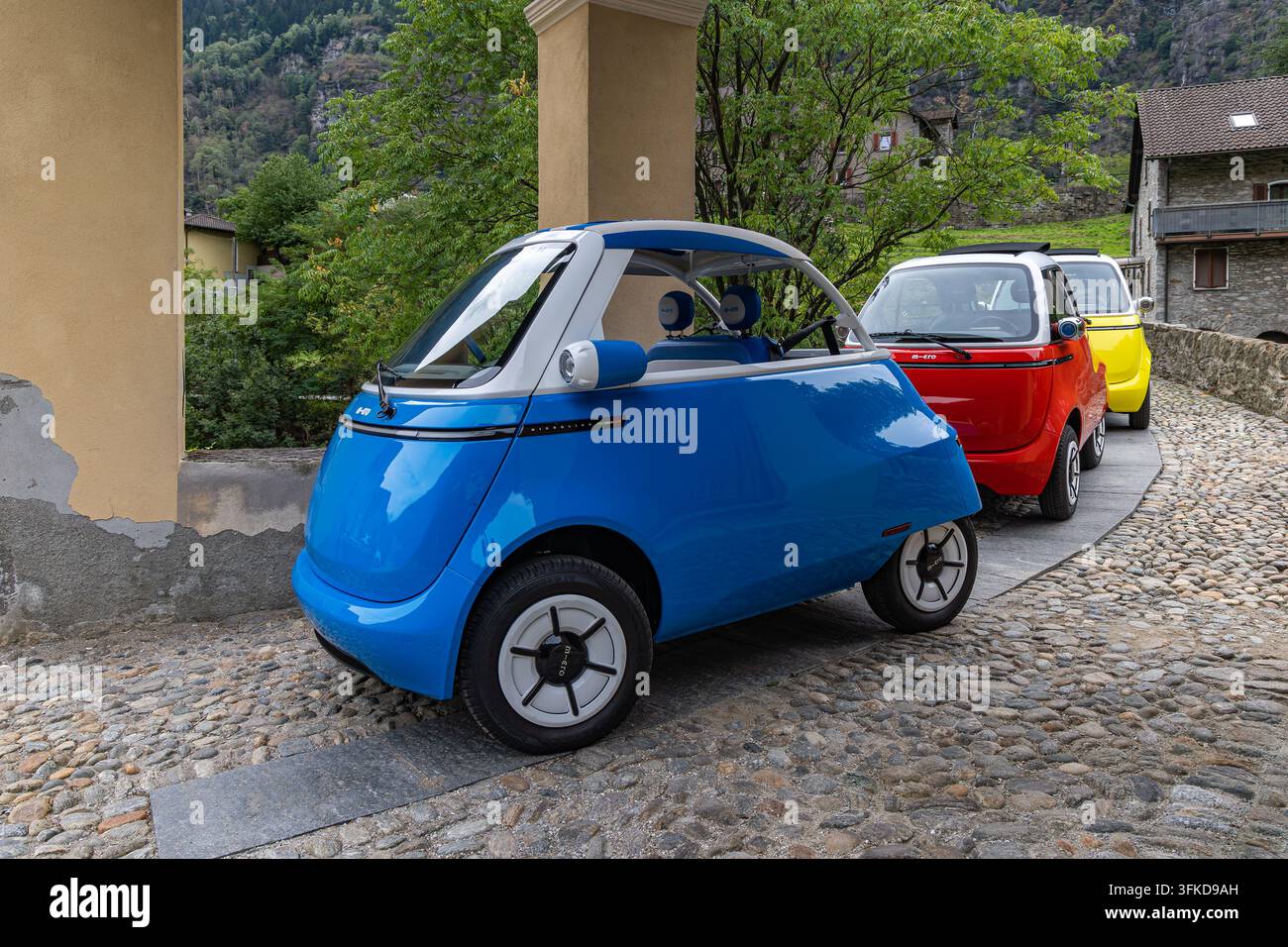 Giornico, Svizzera - 26 agosto 2026: Tre colorati mini auto elettriche Microlino sono esposti sul vecchio ponte di pietra Ai Pontin nel villaggio di Gio Foto Stock