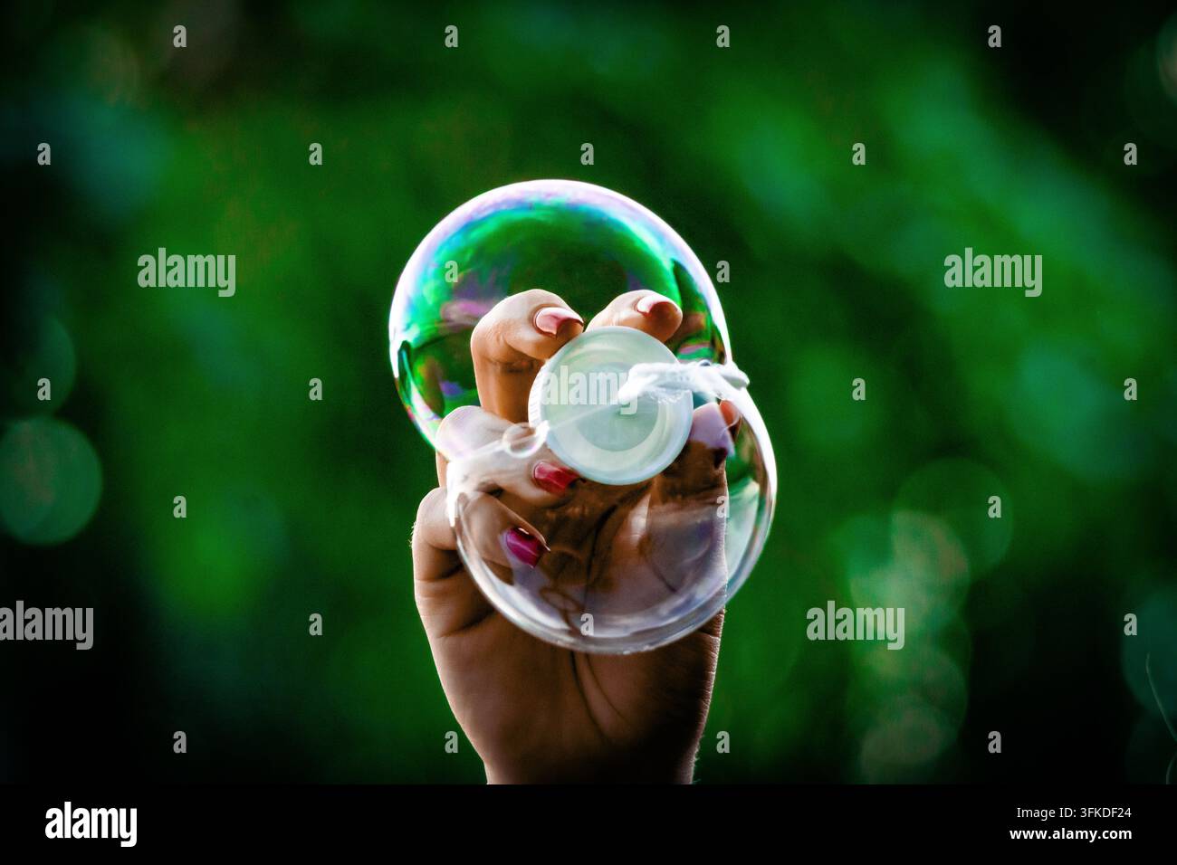 Una mano con unghie rosso vivo solleva una bacchetta per bolle mentre una sfera trasparente si forma contro un bokeh verde lussureggiante. Luce giocosa e dettagli nitidi celebrano una m spensierata Foto Stock