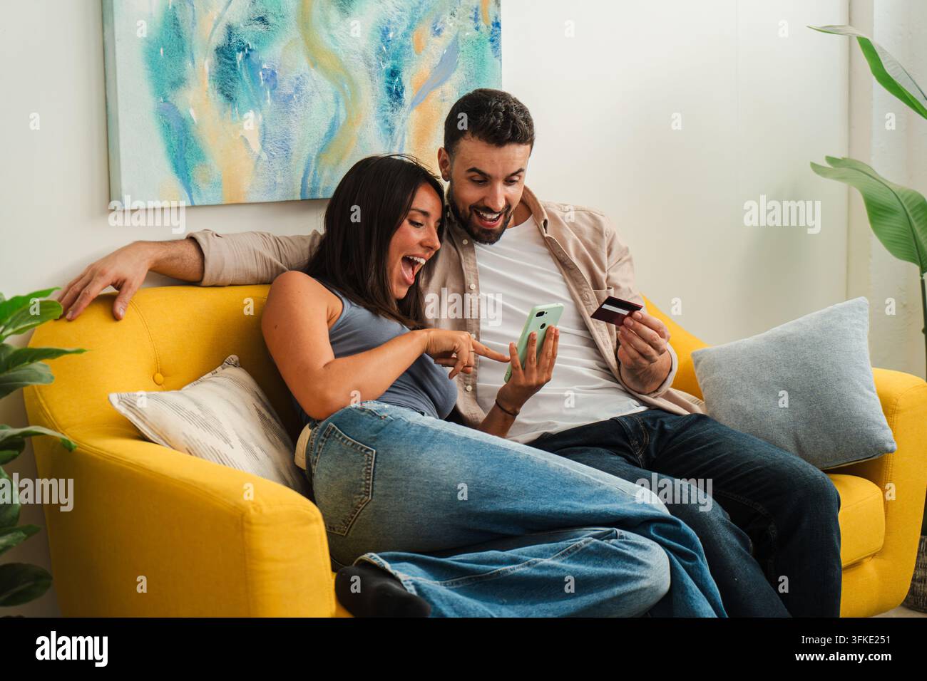 Una coppia si rilassa su un divano giallo brillante, scorrendo con eccitazione sui loro telefoni e condividendo un momento di shopping online. La scena cattura Foto Stock