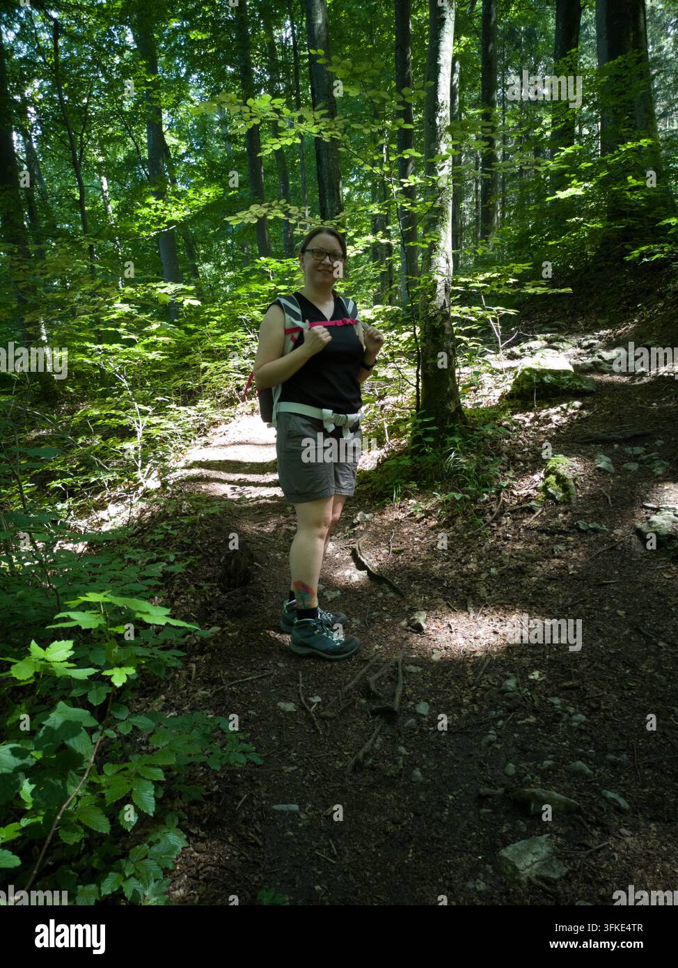 Una donna con occhiali e uno zaino in piedi su un sentiero escursionistico sterrato in una foresta rigogliosa illuminata dal sole. Indossa abiti casual da escursione e scarponi da trekking Foto Stock
