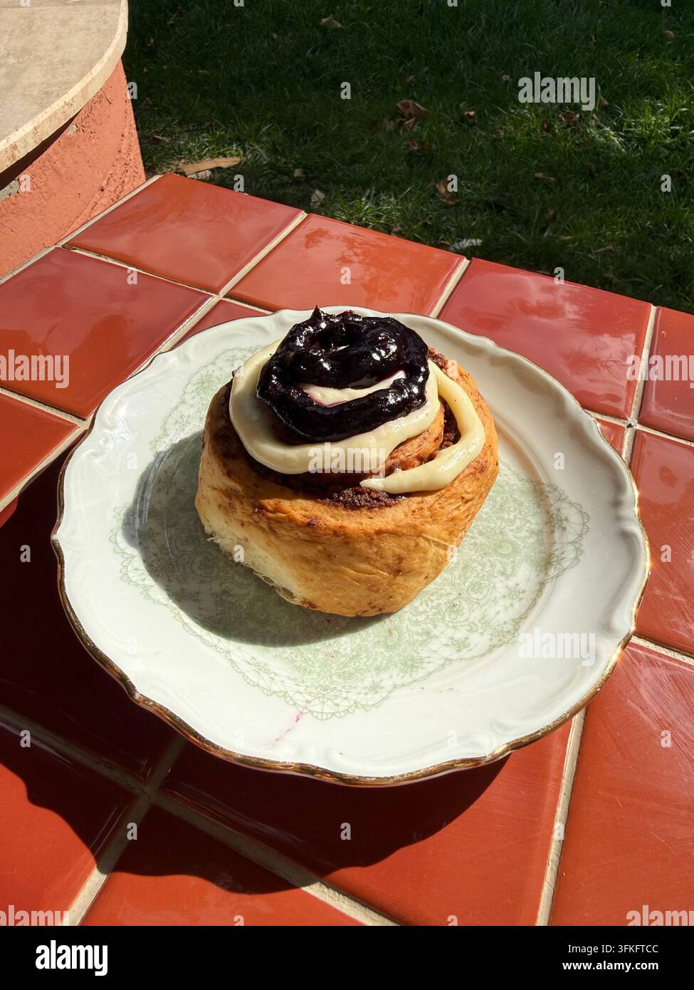 Panino alla cannella appena sfornato ricoperto di glassa al formaggio fresco liscia e ricchezza di compota di frutta, servito su un piatto vintage all'aperto con luce naturale calda. Foto Stock