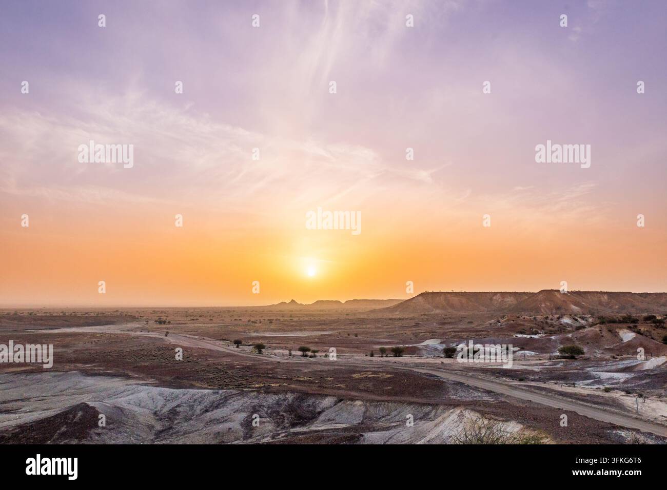 Il sole che tramonta nel cielo nebbioso sopra le badlands erose, le mesas e un sentiero sterrato nel deserto dipinto dell'Outback Foto Stock