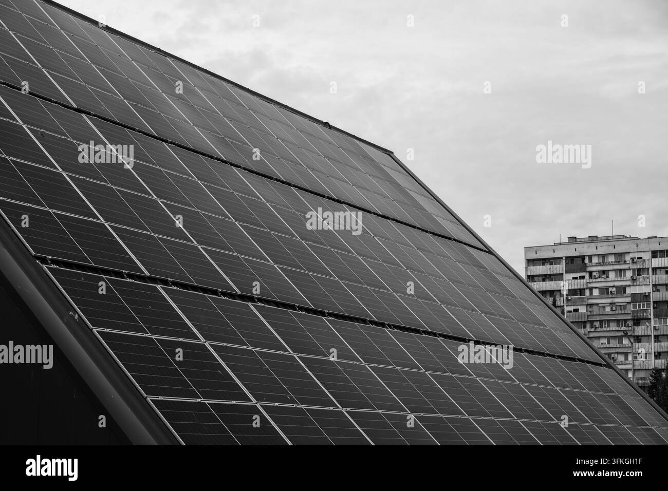 Uno scatto in bianco e nero, con prospettiva a bassa angolazione, di una grande disposizione inclinata di pannelli solari fotovoltaici su un tetto con un condominio a più piani sullo sfondo Foto Stock
