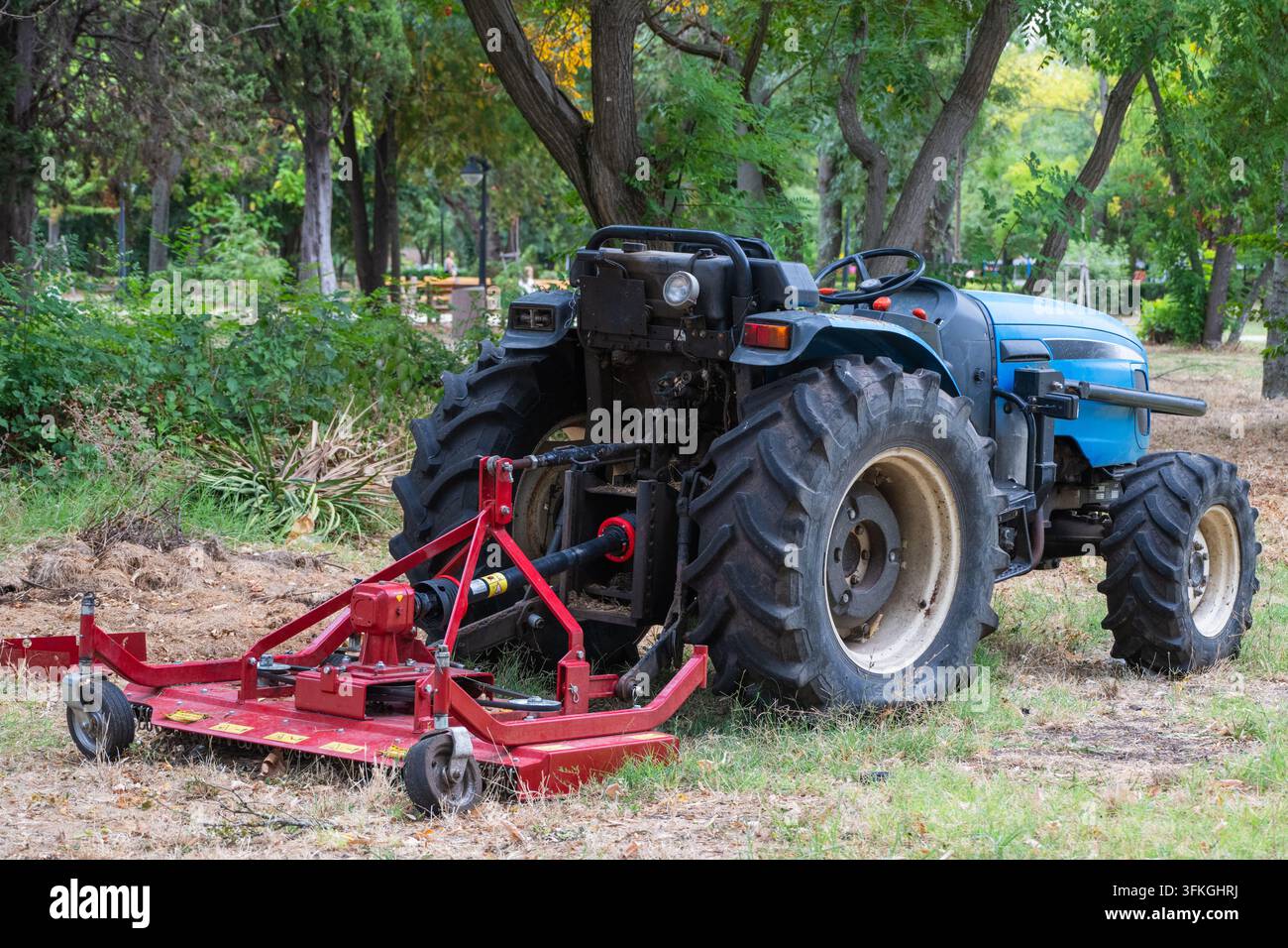 Un trattore compatto blu brillante equipaggiato con un accessorio rosso per falciatrice a spandau posteriore parcheggiato in un parco erboso circondato da alberi. Foto Stock
