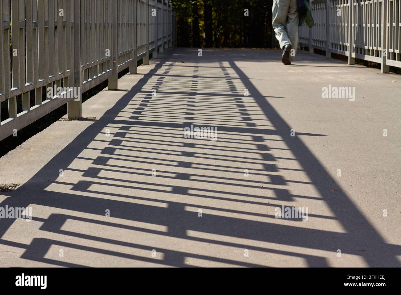 Ombre geometriche da ringhiere metalliche si estendono su un ponte di cemento mentre una persona parzialmente visibile si allontana verso gli alberi alla luce del sole Foto Stock