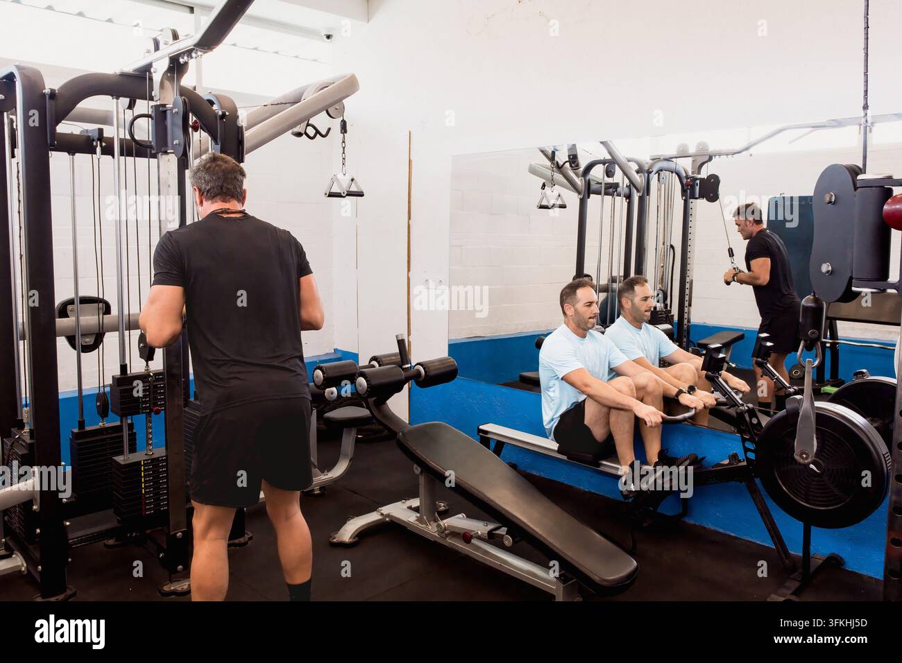 Vista posteriore di un allenatore atletico in nero che osserva un uomo in blu che utilizza un vogatore seduto durante una sessione di allenamento in palestra. Foto Stock