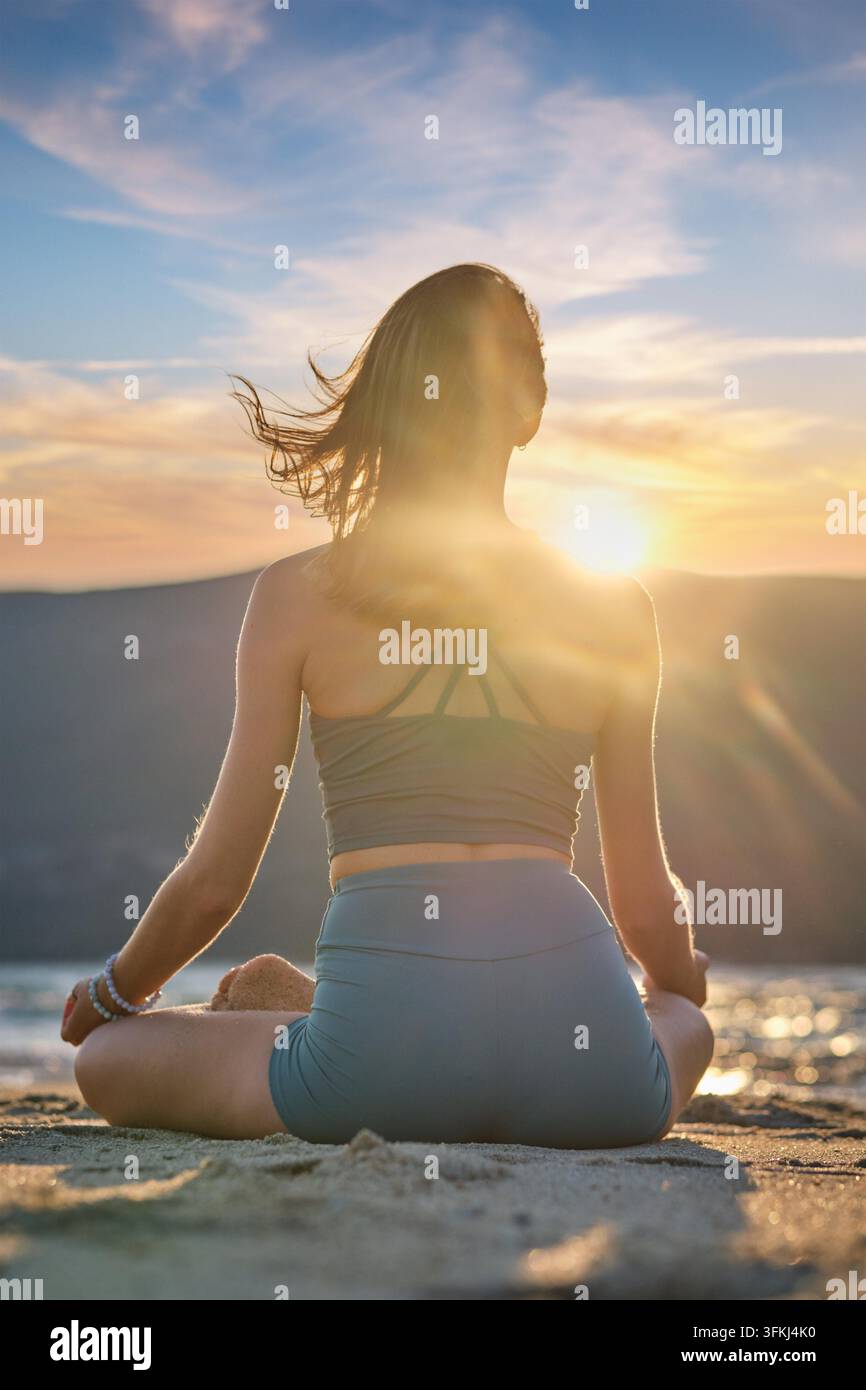 Una donna siede a Padmasana sulla spiaggia, meditando con il Chin mudra mentre il sole tramonta, creando un'atmosfera serena e rilassante. Foto Stock