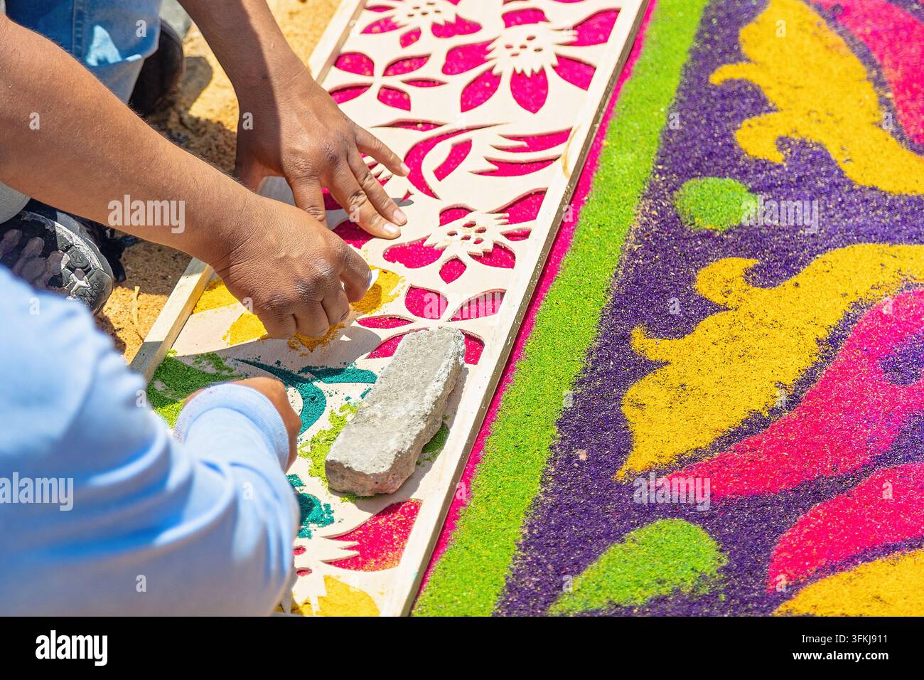 Primo piano di mani che usano uno stencil floreale e segatura colorata per creare una tradizionale alfombra della Settimana Santa. Foto Stock