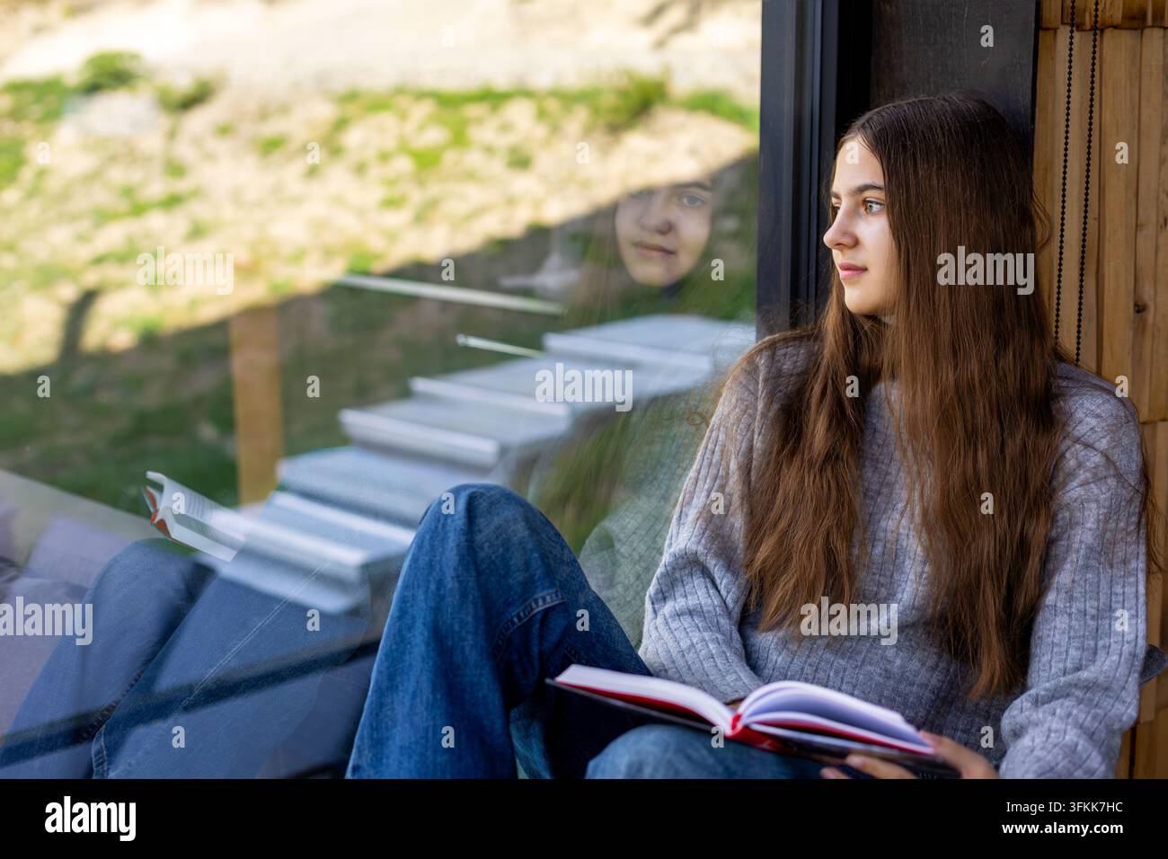 Giovane donna che legge vicino a una finestra con riflesso. Giovane donna seduta vicino a una grande finestra tiene un libro aperto e guarda fuori, con il suo riflesso visibile Foto Stock