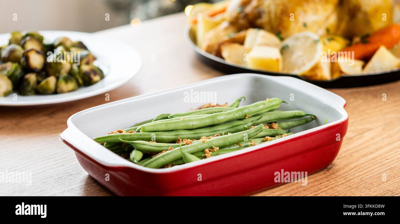 Fagiolini verdi freschi sormontati da cipolle fritte croccanti serviti in una pirofila rossa, con tacchino arrosto, verdure e cavolfiori sullo sfondo. Foto Stock