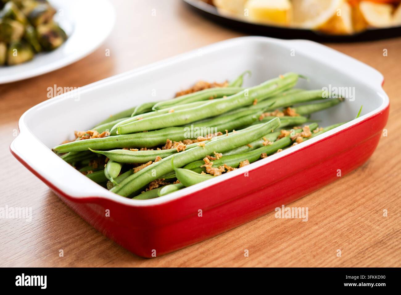 Fagiolini verdi freschi sormontati da cipolle fritte croccanti serviti in una pirofila rossa, con tacchino arrosto, verdure e cavolfiori sullo sfondo. Foto Stock