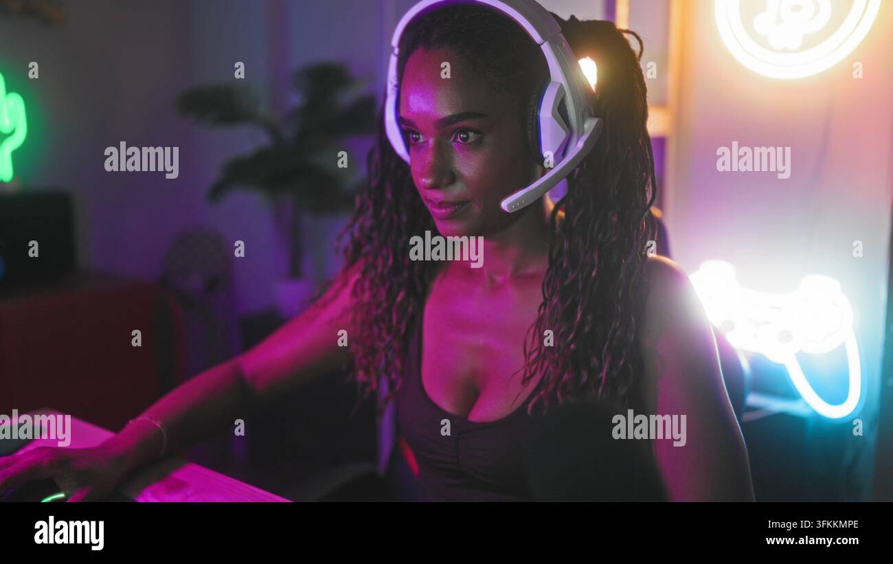 Donna con auricolari che gioca a una scrivania da studio con controller neon e braccia incrociate, sguardo concentrato; concentrazione determinata. Foto Stock