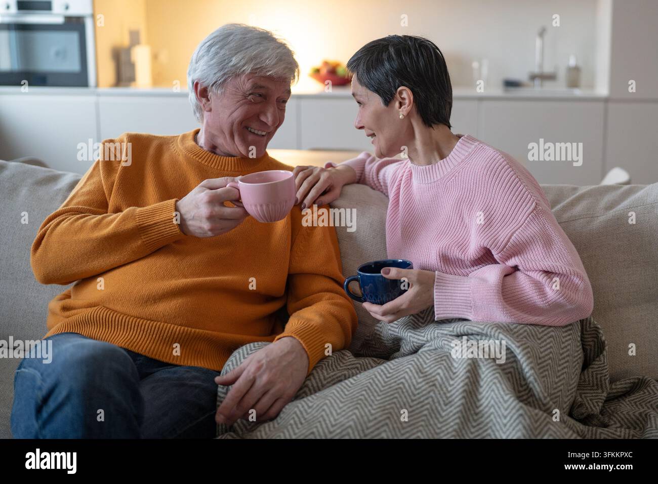 Una coppia di anziani affettuosi beve il tè e si sorride, seduti insieme sul divano a casa Foto Stock