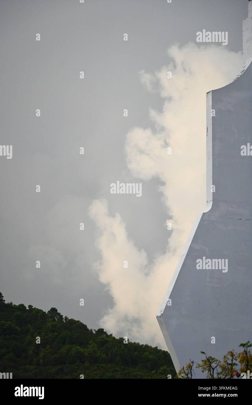 Fotografia verticale dettagliata di nuvole bianche che si alzano dietro una facciata di edificio moderno e un paesaggio collinare verde Foto Stock