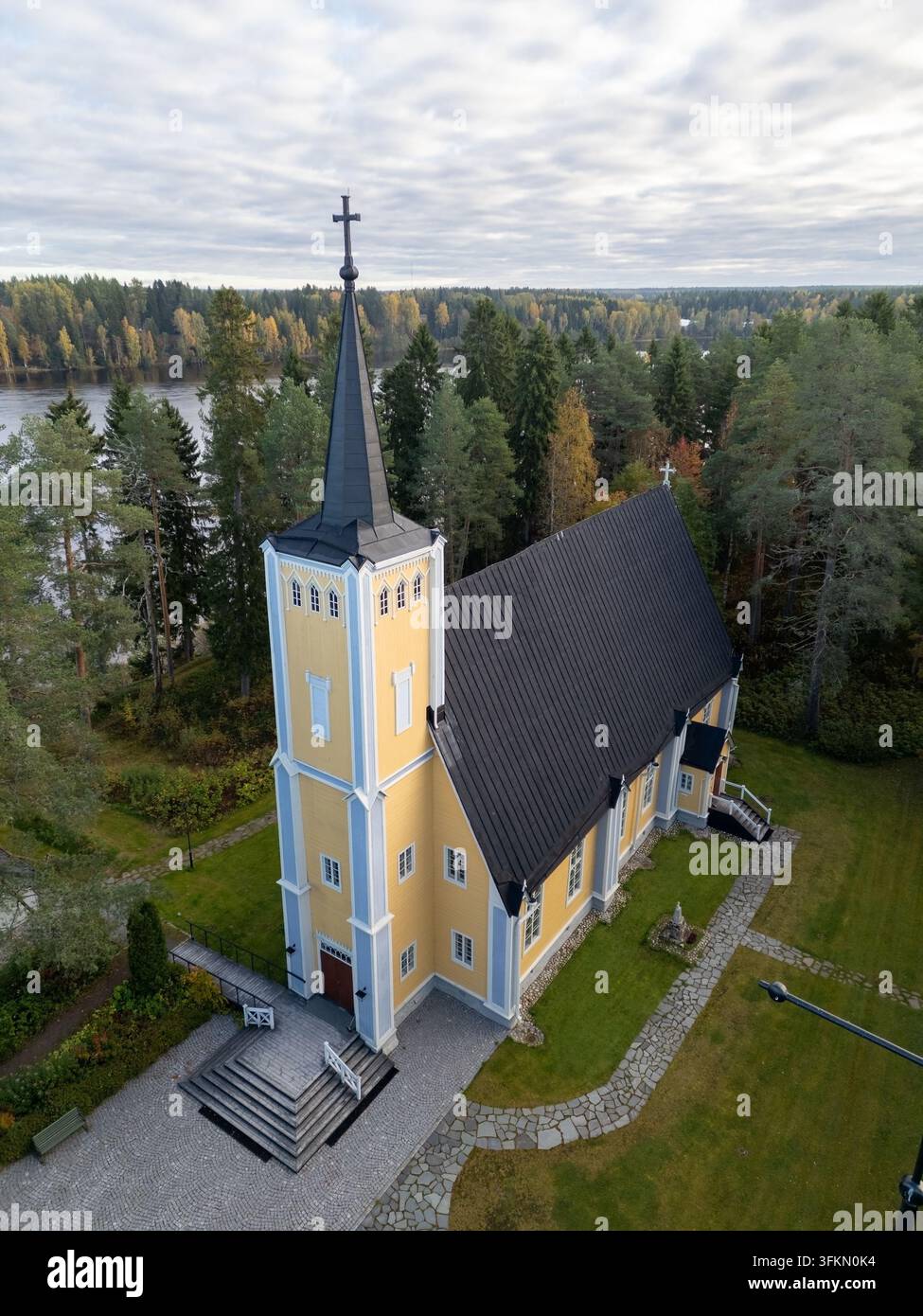 Vista aerea della storica chiesa di Muhos con le sue pareti in legno giallo e il tetto nero, circondata da foreste di pini e fogliame autunnale vicino al fiume Foto Stock