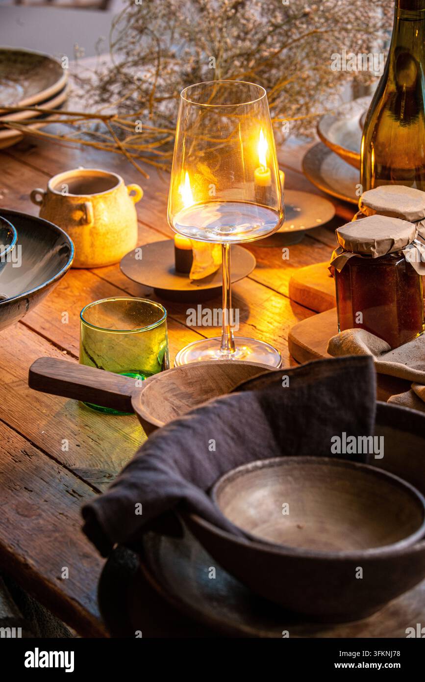 Impostazione rustica calda del tavolo con un bicchiere di vino trasparente, un tumbler verde, atmosfera illuminata a candela, ciotole di legno e barattoli di miele. Foto Stock