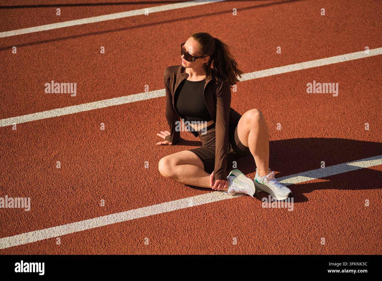 Una sportiva moderna gira la testa mentre si riposa sulla pista tra le linee bianche della pista da corsa all'aperto Foto Stock
