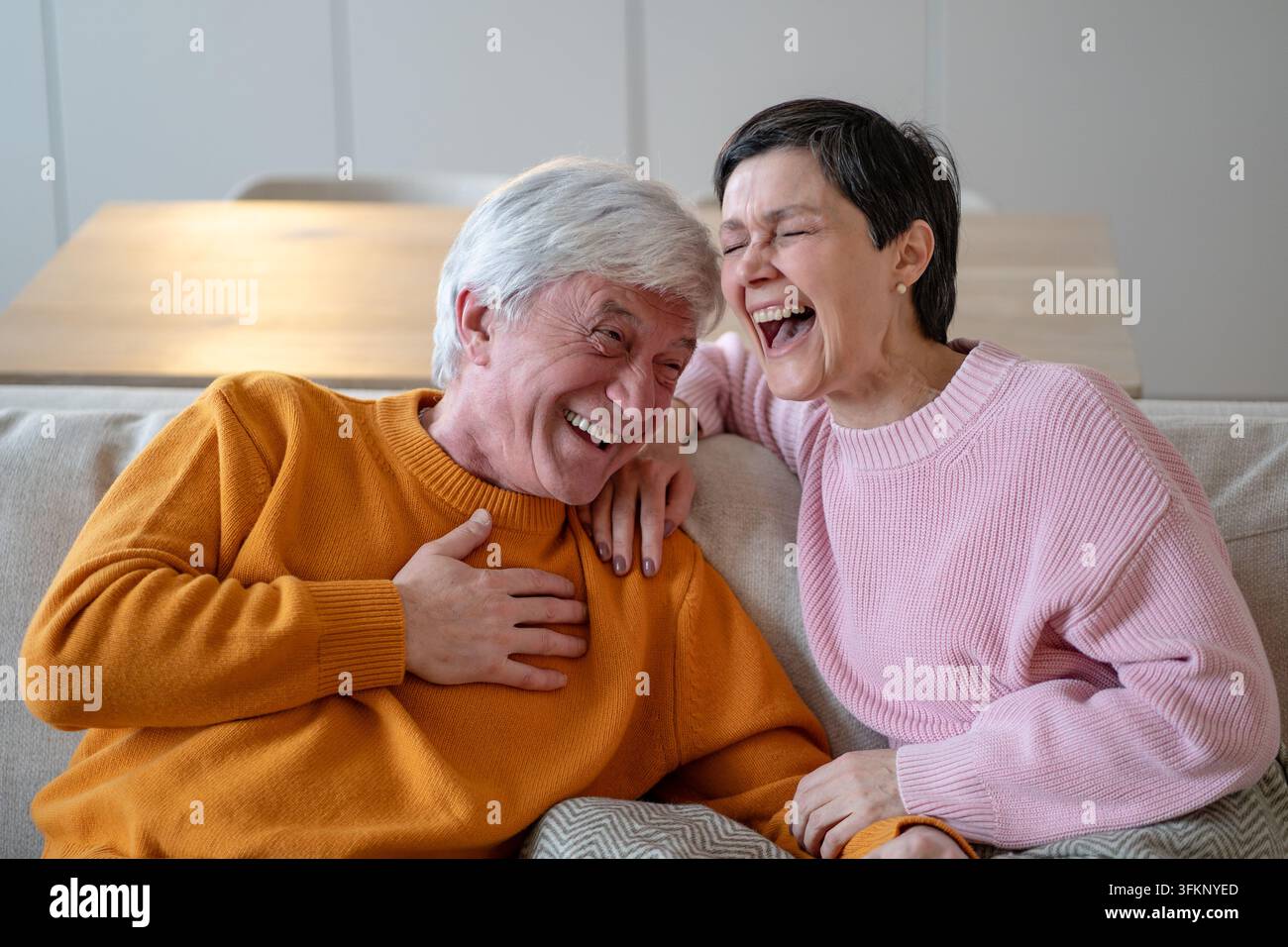 Coniugi anziani gioiosi si abbracciano e ridono insieme, seduti vicini su un comodo divano a casa Foto Stock