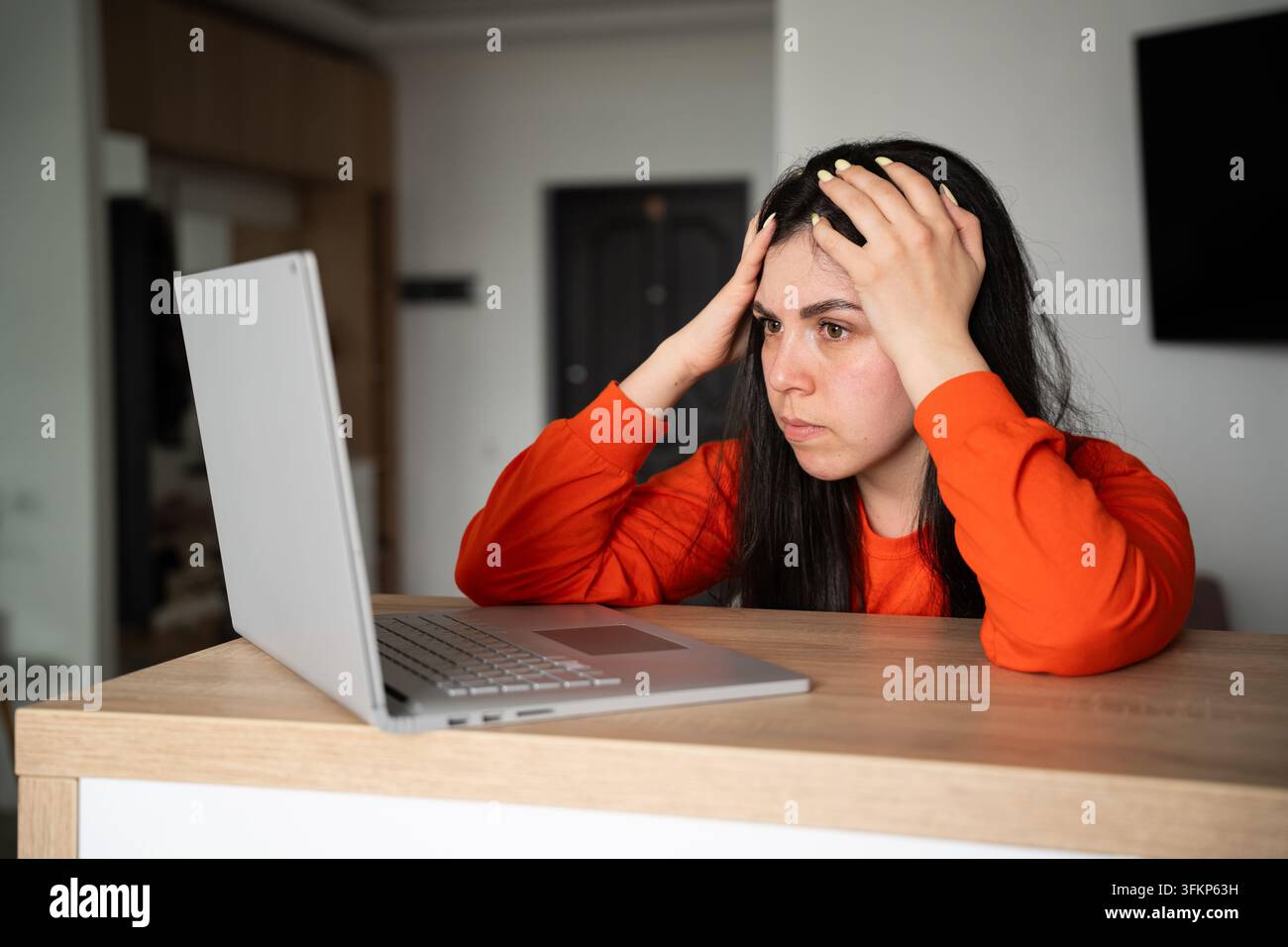 Donna frustrata che si tiene la testa mentre usa un laptop alla scrivania in un ufficio domestico. Stress sul lavoro, frustrazione tecnologica e ansia per le scadenze. Foto Stock