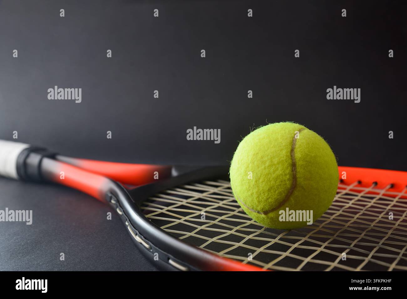 Una palla da tennis gialla poggia sulle corde di una racchetta rossa e nera, con un'altra racchetta dietro su uno sfondo scuro da studio. Foto Stock