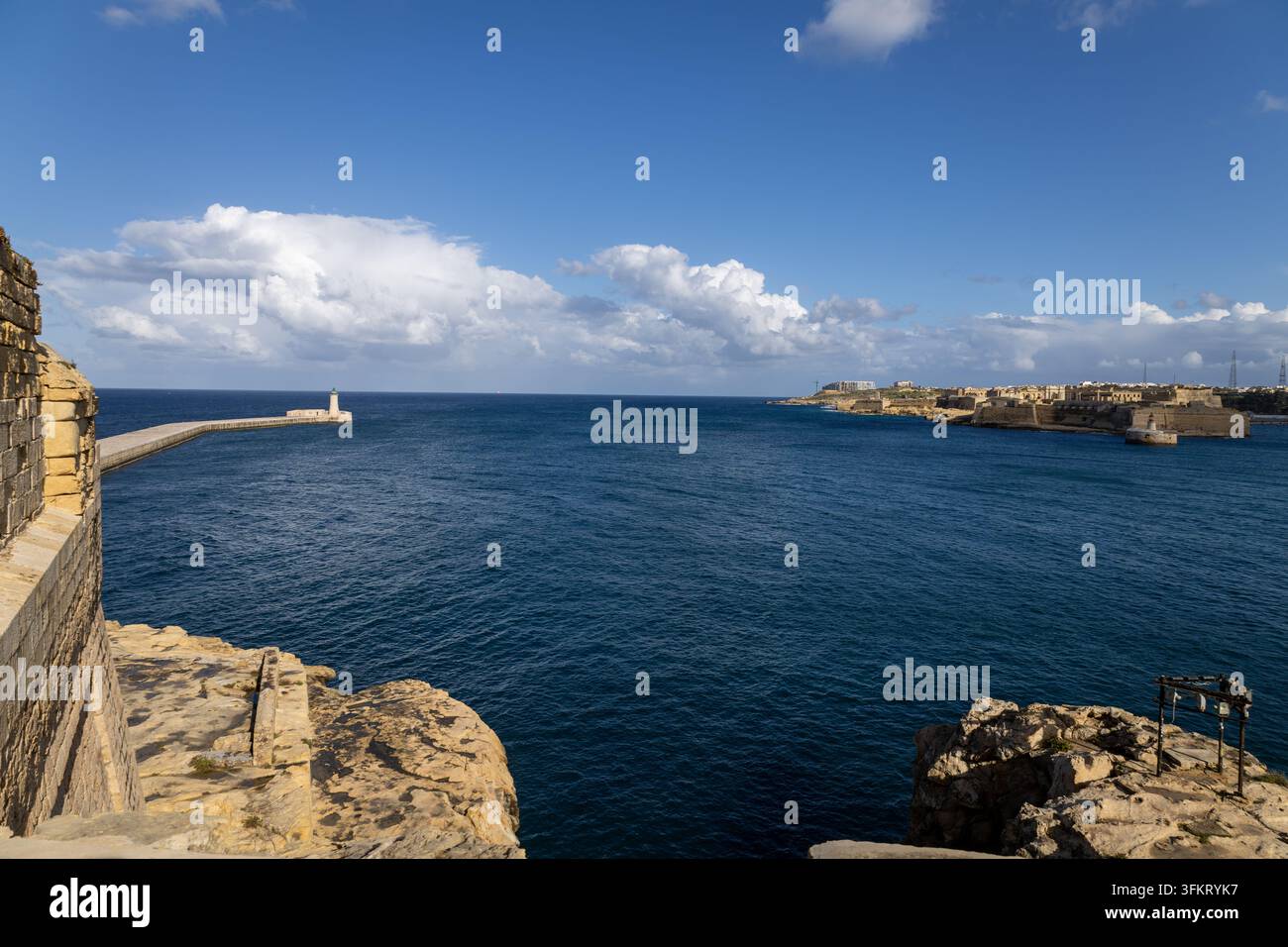 Vista panoramica da Fort St. Elmo sul mare blu, fortificazioni storiche in calcare, strutture portuali, un faro e edifici urbani distanti benea Foto Stock