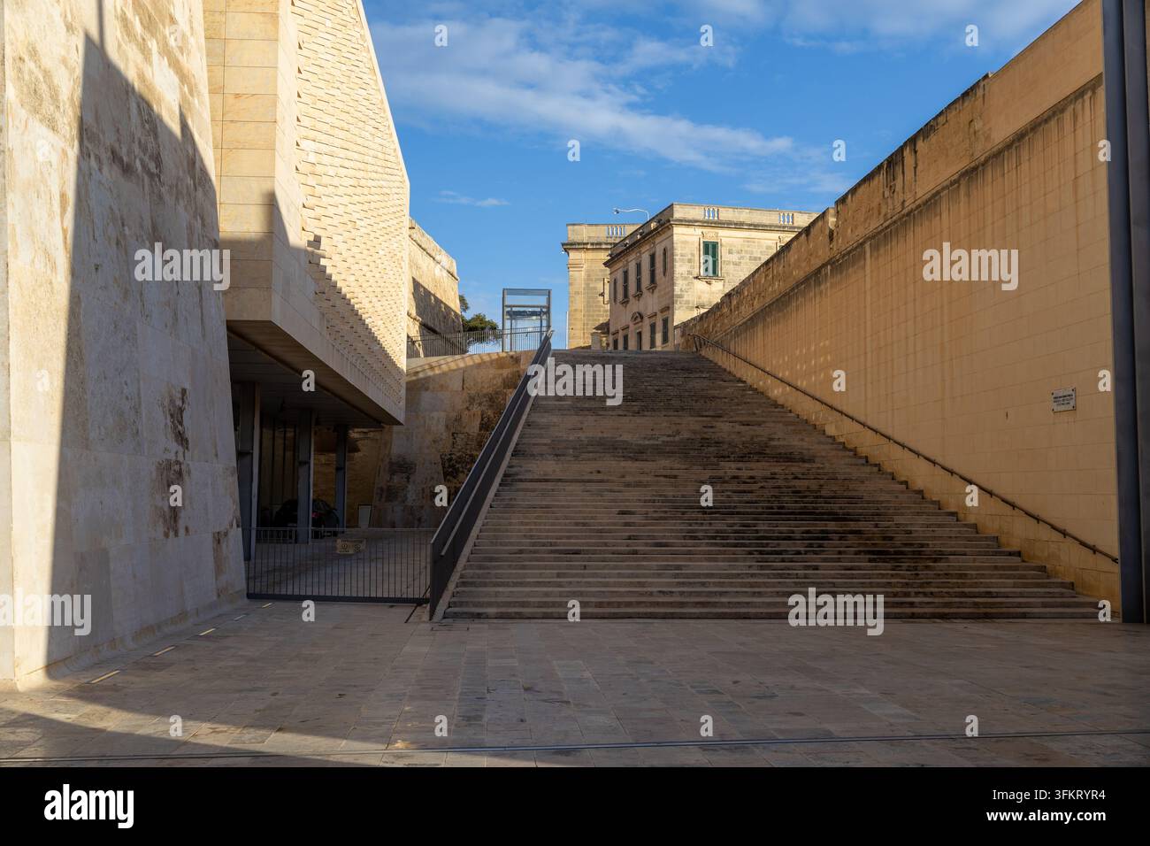 Muri di calcare illuminati dal sole, una larga scala, ringhiere e forme architettoniche angolari sotto un cielo blu terso a La Valletta, Malta. Foto Stock