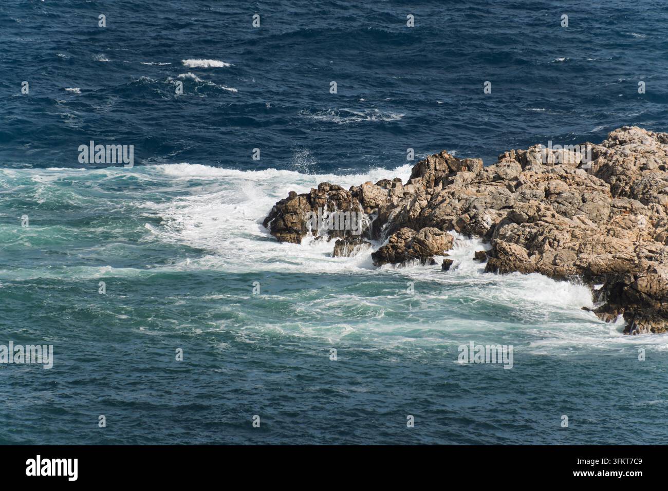Onde blu profonde che avvolgono un punto roccioso di calcare pallido con schiuma bianca turbinante. Foto Stock