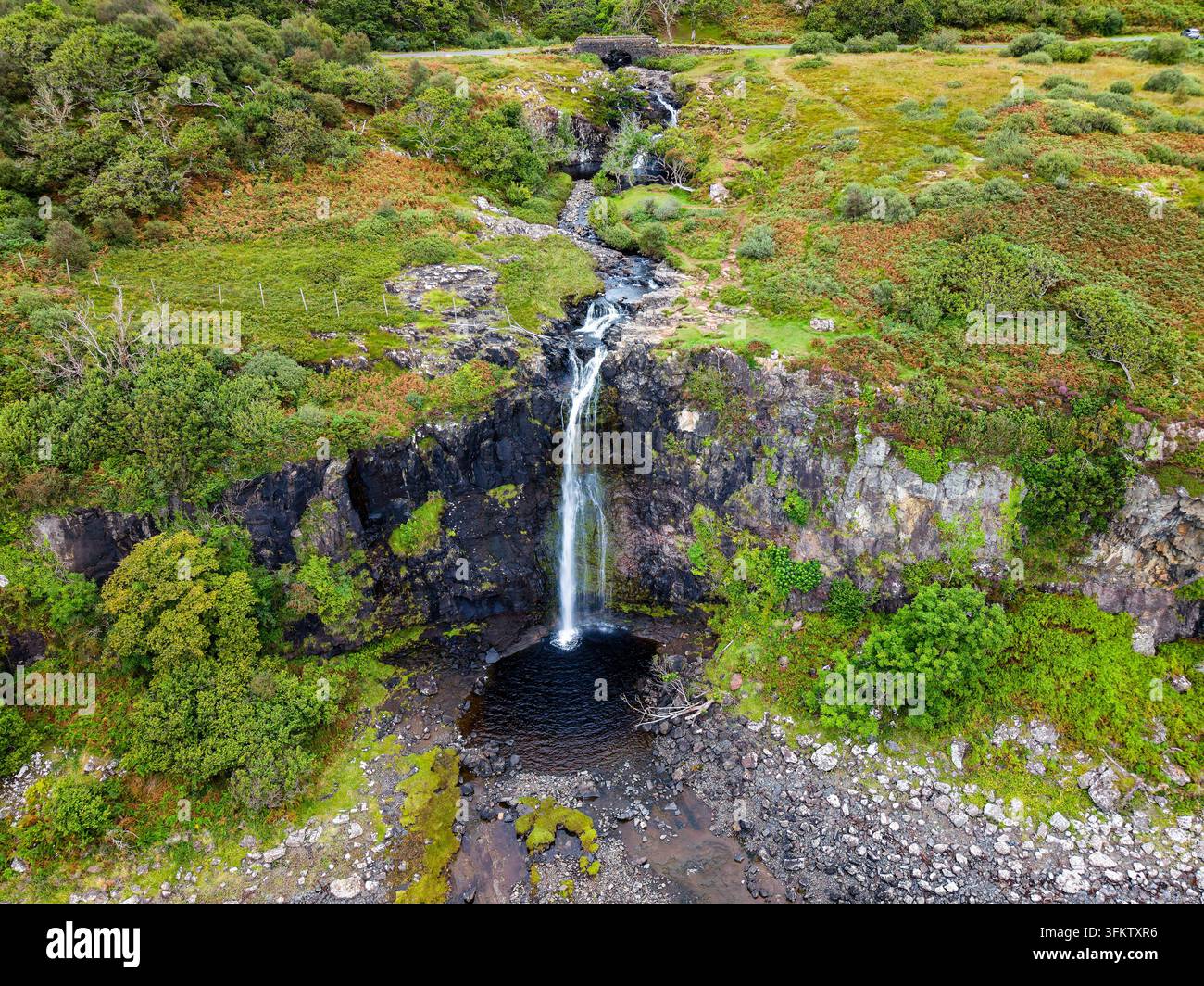 Vista aerea di una cascata che cade su una parete rocciosa scura verso la riva rocciosa, torrente costiero che drena il terreno dell'altopiano attraverso pendici verdeggianti Foto Stock