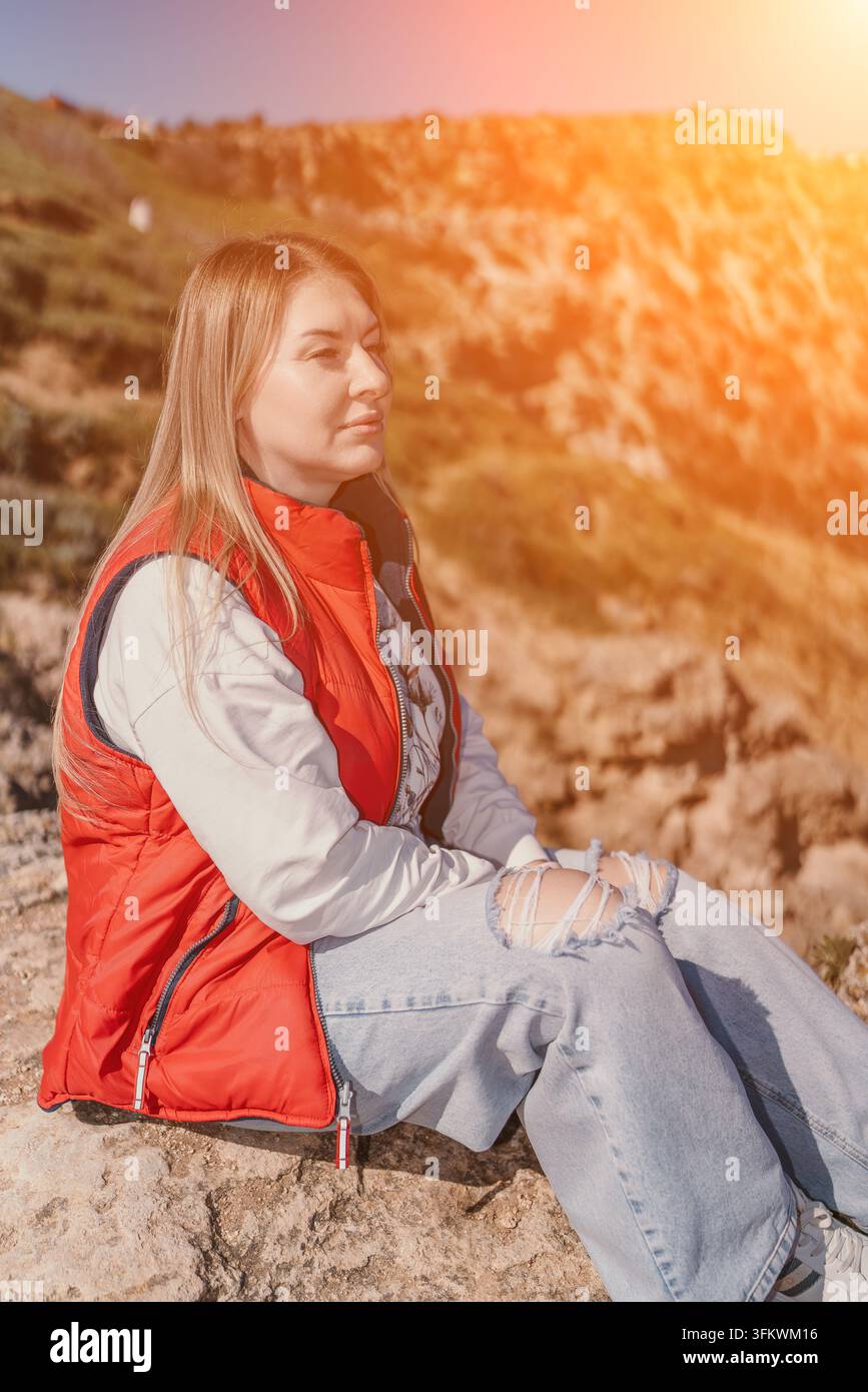 Donna seduta su una scogliera rocciosa che indossa un giubbotto rosso e jeans strappati, godendosi la calda luce del tramonto all'aperto Foto Stock