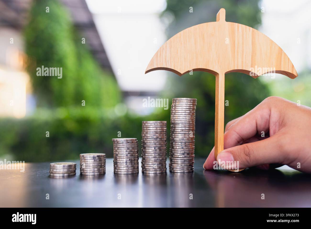 Concetto di assicurazione finanziaria e protezione del risparmio, mano che tiene un ombrello di legno sopra pile di monete ascendenti sul tavolo, simboleggiando la salvaguardia della ricchezza Foto Stock