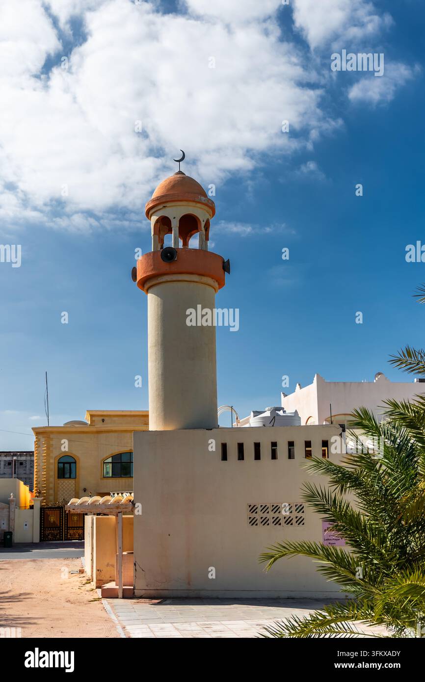 Minareto tradizionale di una moschea con una cupola arancione che si innalza sopra muri bianchi e palme sotto un cielo blu brillante con nuvole delicate. Foto Stock