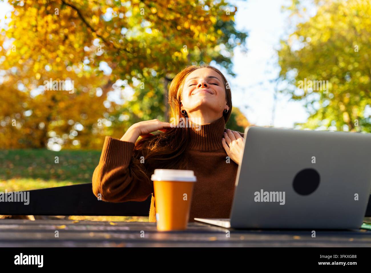 Donna con laptop e caffè da asporto che gode del tempo soleggiato autunnale nel parco cittadino Foto Stock