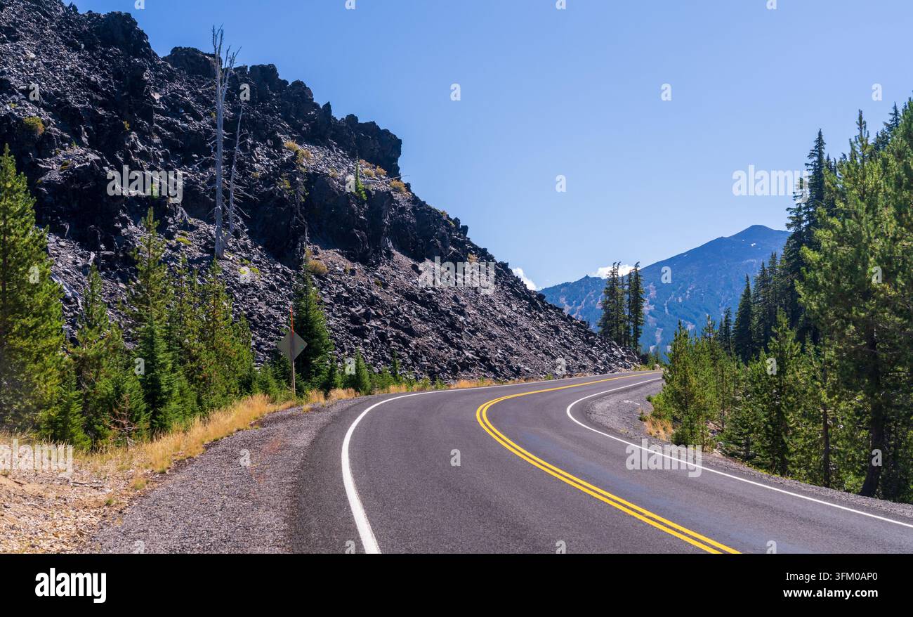 Una strada asfaltata sinuosa si snoda oltre un ripido argine di flusso di lava di basalto scuro e alberi sempreverdi nel deserto dell'Oregon. Foto Stock