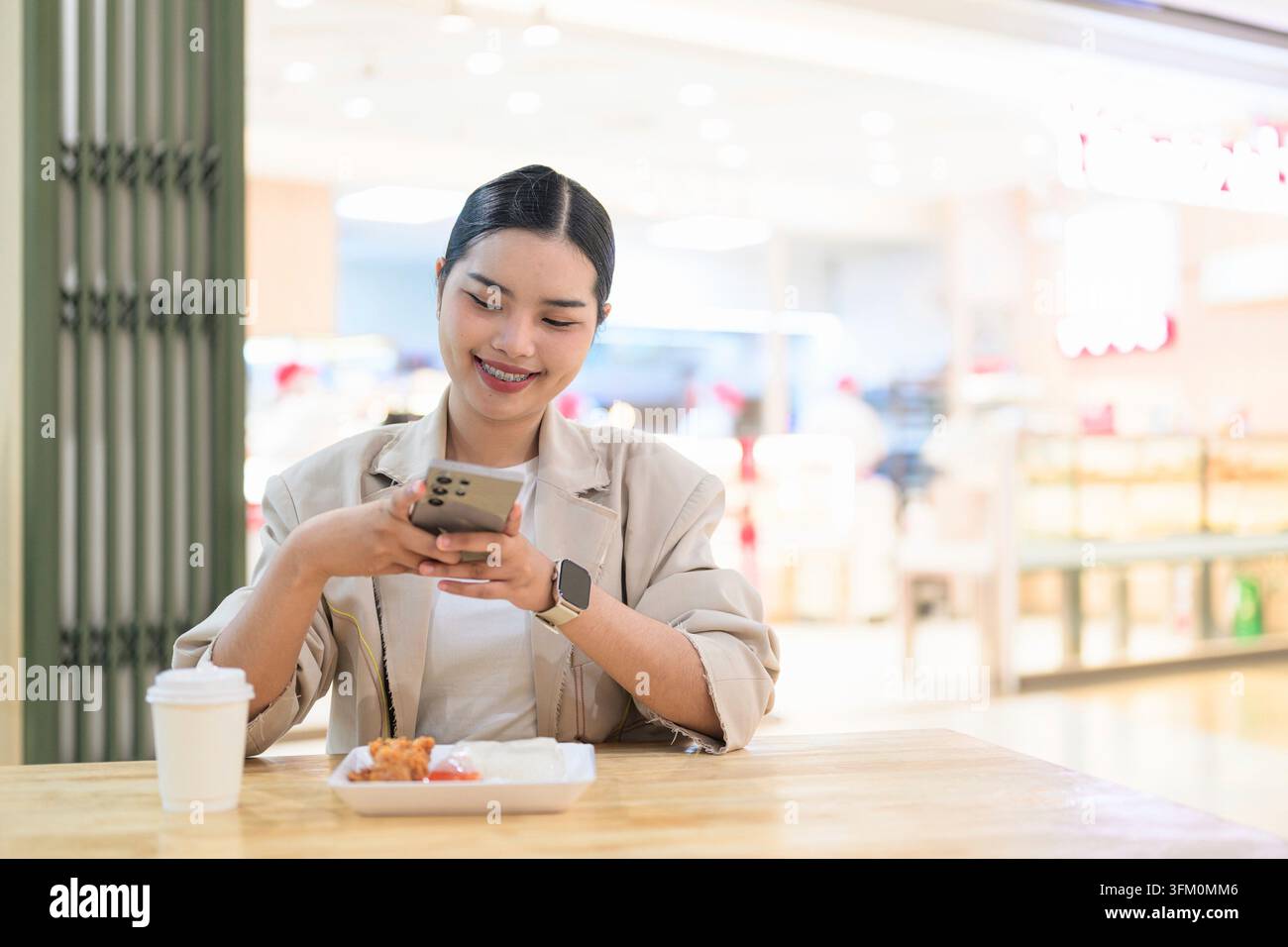 Una giovane donna d'affari thailandese del sud-est asiatico, che guarda in basso e sorride mentre tiene in mano e utilizza uno smartphone. Sta indossando un Foto Stock