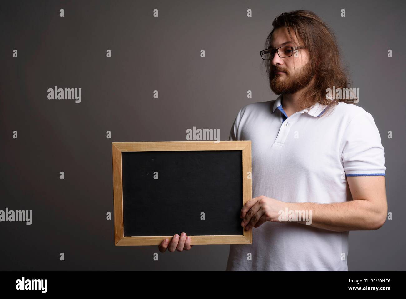 Ritratto in studio di un giovane uomo caucasico pensieroso con capelli lunghi che tiene una lavagna vuota. Ideale per messaggi personalizzati, idee visionarie e pubblicità Foto Stock