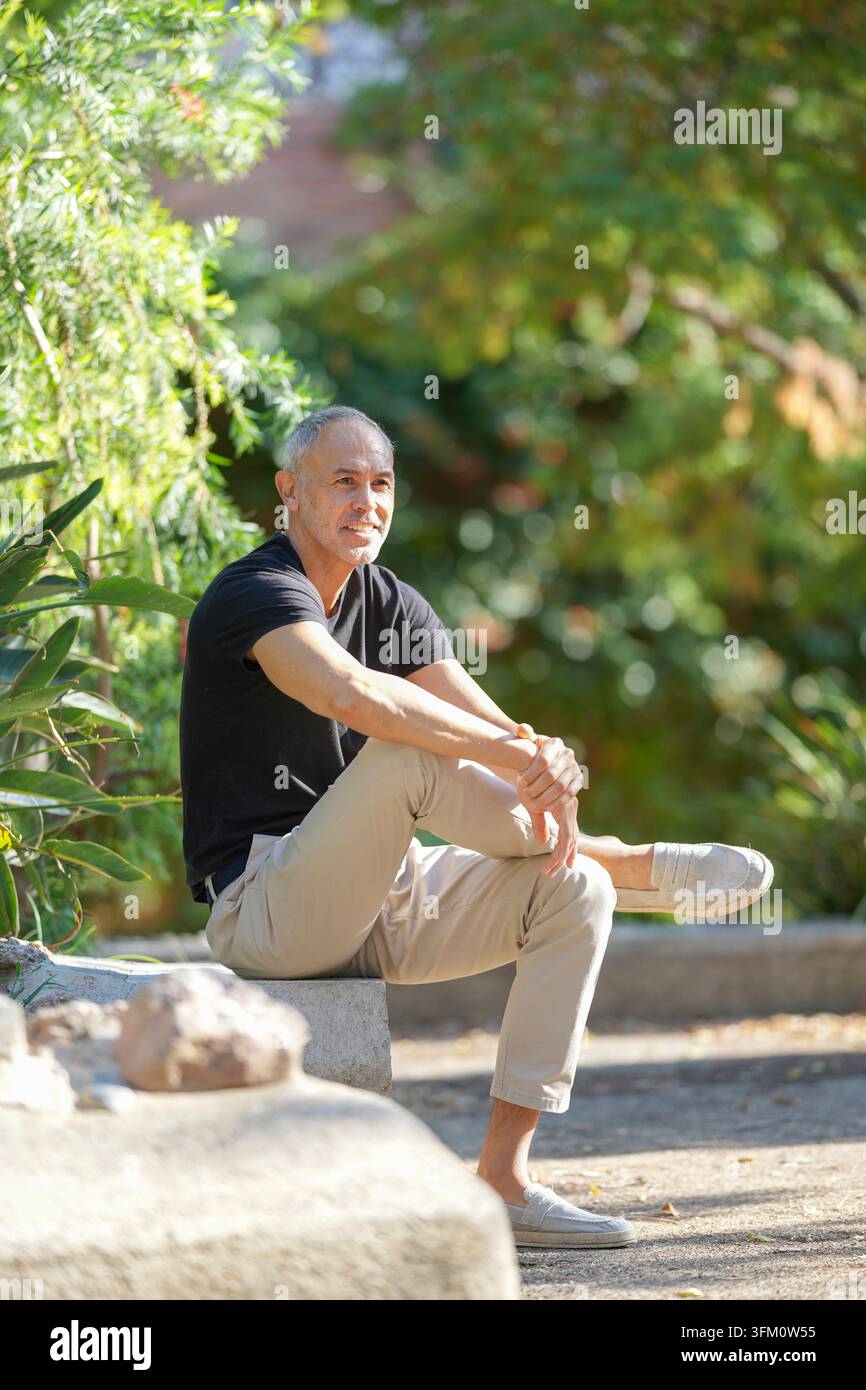 Un uomo con una camicia nera e pantaloni abbronzati siede su una panchina in un parco. Sta sorridendo e si sta godendo la vita all'aria aperta Foto Stock