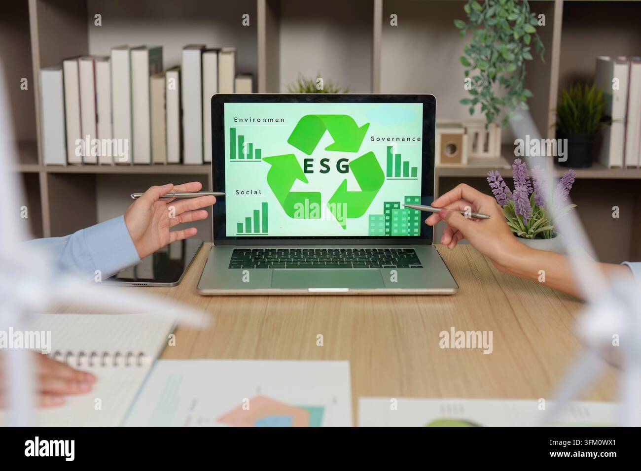Due professionisti aziendali indicano uno schermo di laptop che mostra una grafica ESG con simboli di riciclo e grafici, discutendo la sostenibilità aziendale s Foto Stock
