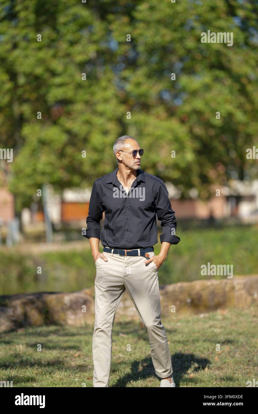 Un uomo con una camicia nera e pantaloni abbronzati si trova in un campo erboso. Indossa gli occhiali da sole e ha le mani nelle tasche. Concetto di relax e le Foto Stock