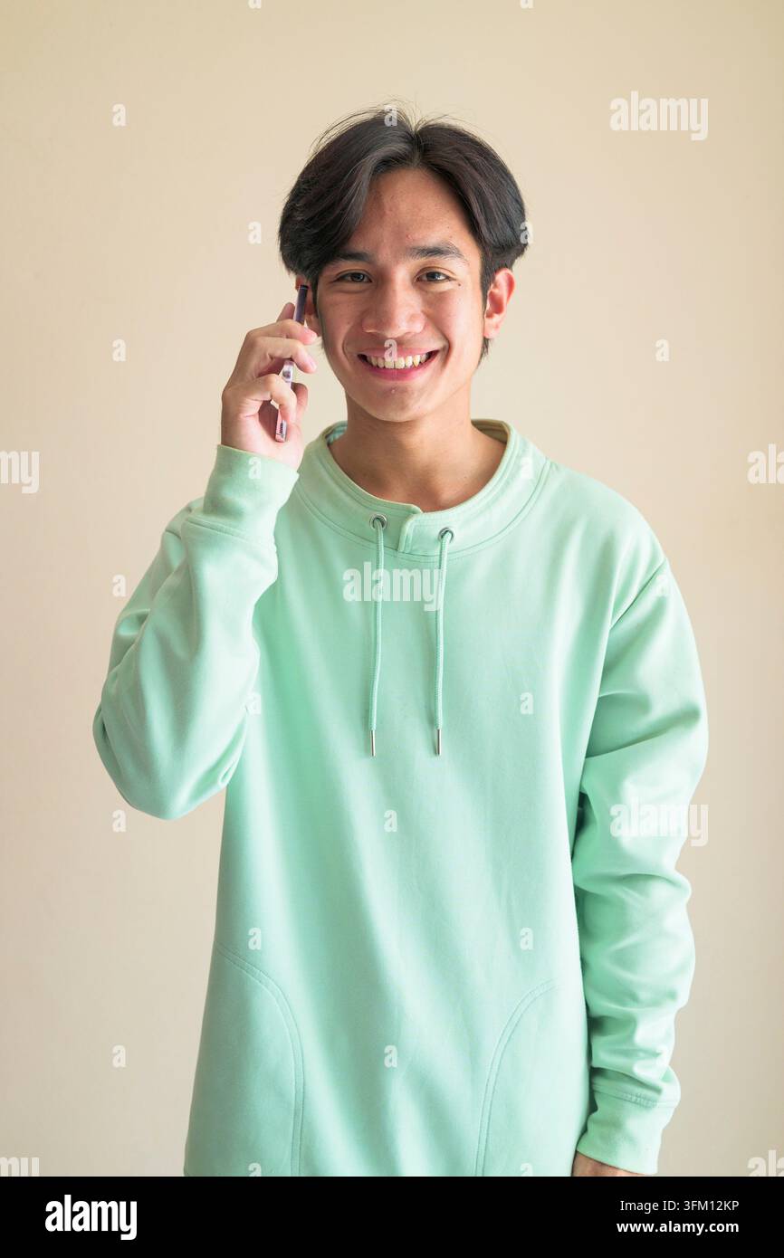Un ritratto in studio di un giovane asiatico allegro della regione di Isan in Tailandia, che tiene un telefono cellulare all'orecchio mentre sorride ampiamente Sembra serio Foto Stock