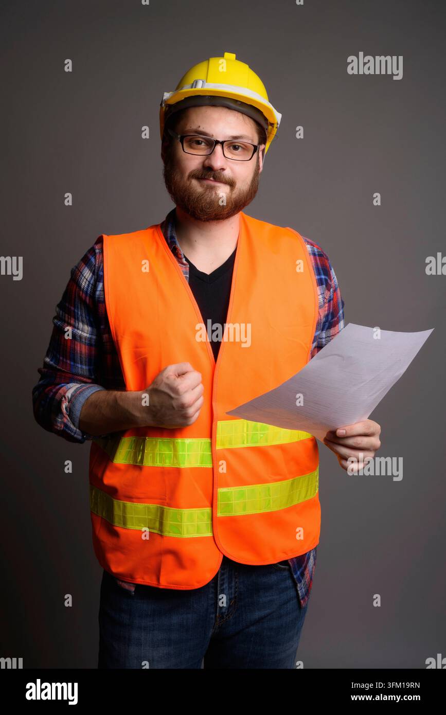 Ritratto a tre quarti di un uomo barbetto felice in un giubbotto di sicurezza e un casco che tiene un documento. Ideale per costruzioni di successo, ingegneria e Foto Stock