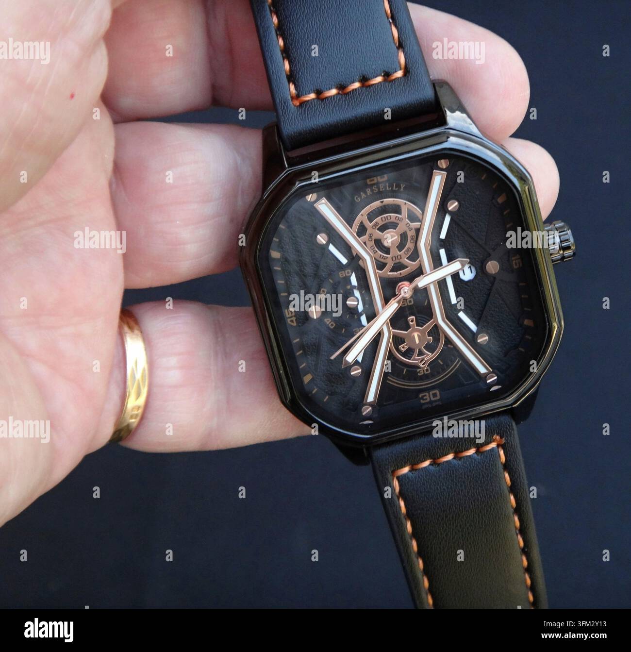 Mano che tiene un orologio da polso maschile nero quadrato con cinturino in pelle cucito arancione contrastante Foto Stock