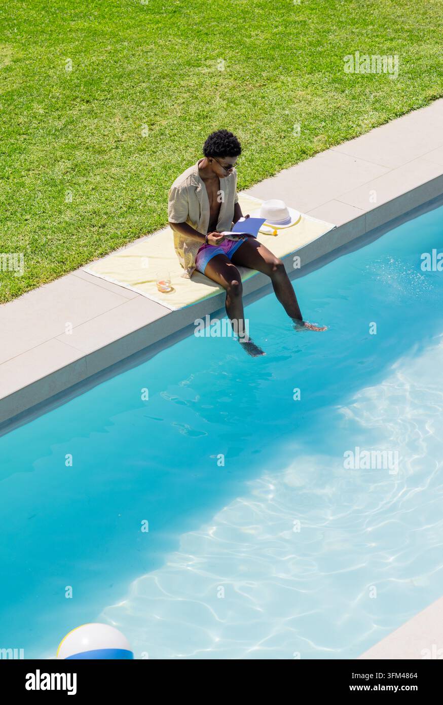 Uomo adulto seduto in piscina, che immerge i piedi, legge su un tablet viola, indossa una camicia e il costume da bagno, pallone da spiaggia. All'aperto, cortile, acqua, luce solare, asciugamano, cappello, occhiali da sole Foto Stock